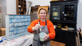 Eine Frau mit Schrze steht in einer Werkstatt und hlt einen Farbtopf